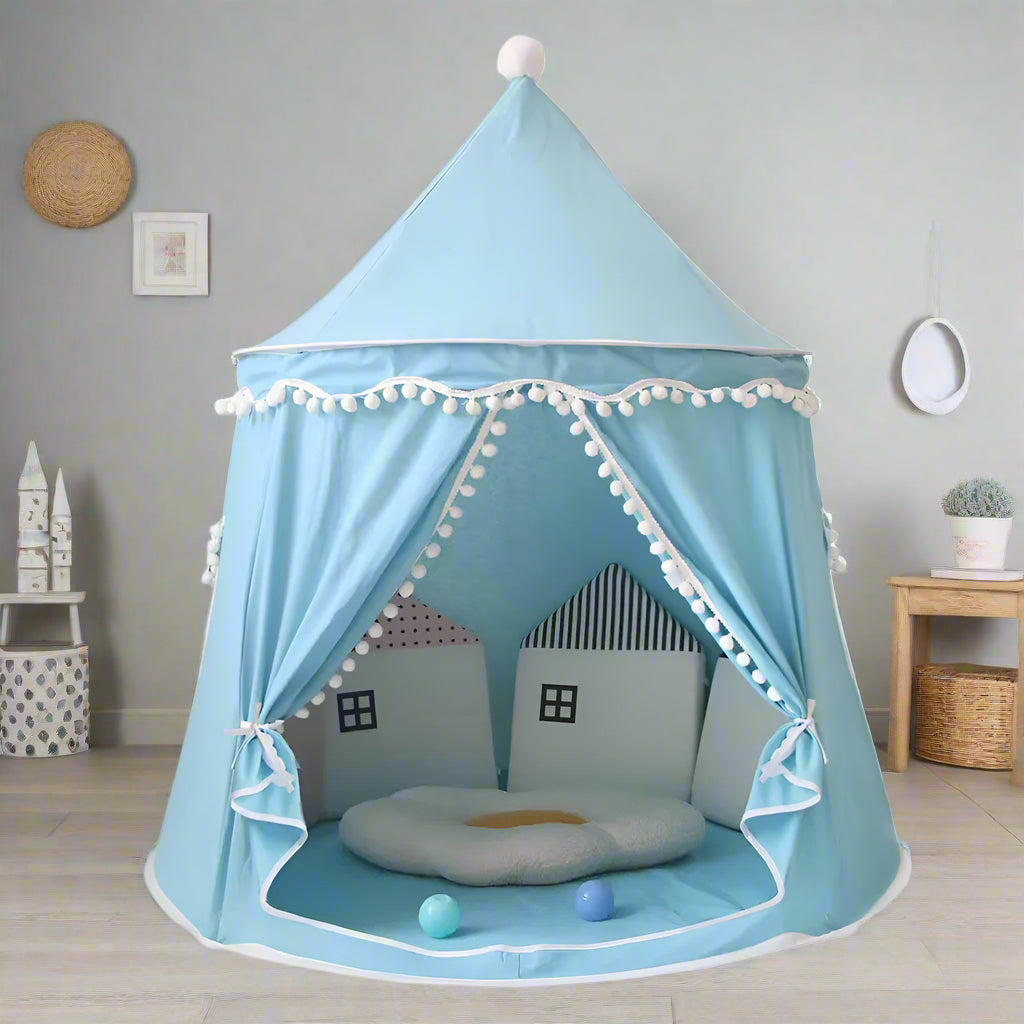 A beige Children's Bali Teepee Tent with a whimsical design, featuring pom-pom details and a small house shape on the front. The tent is set up indoors on a white carpet.
