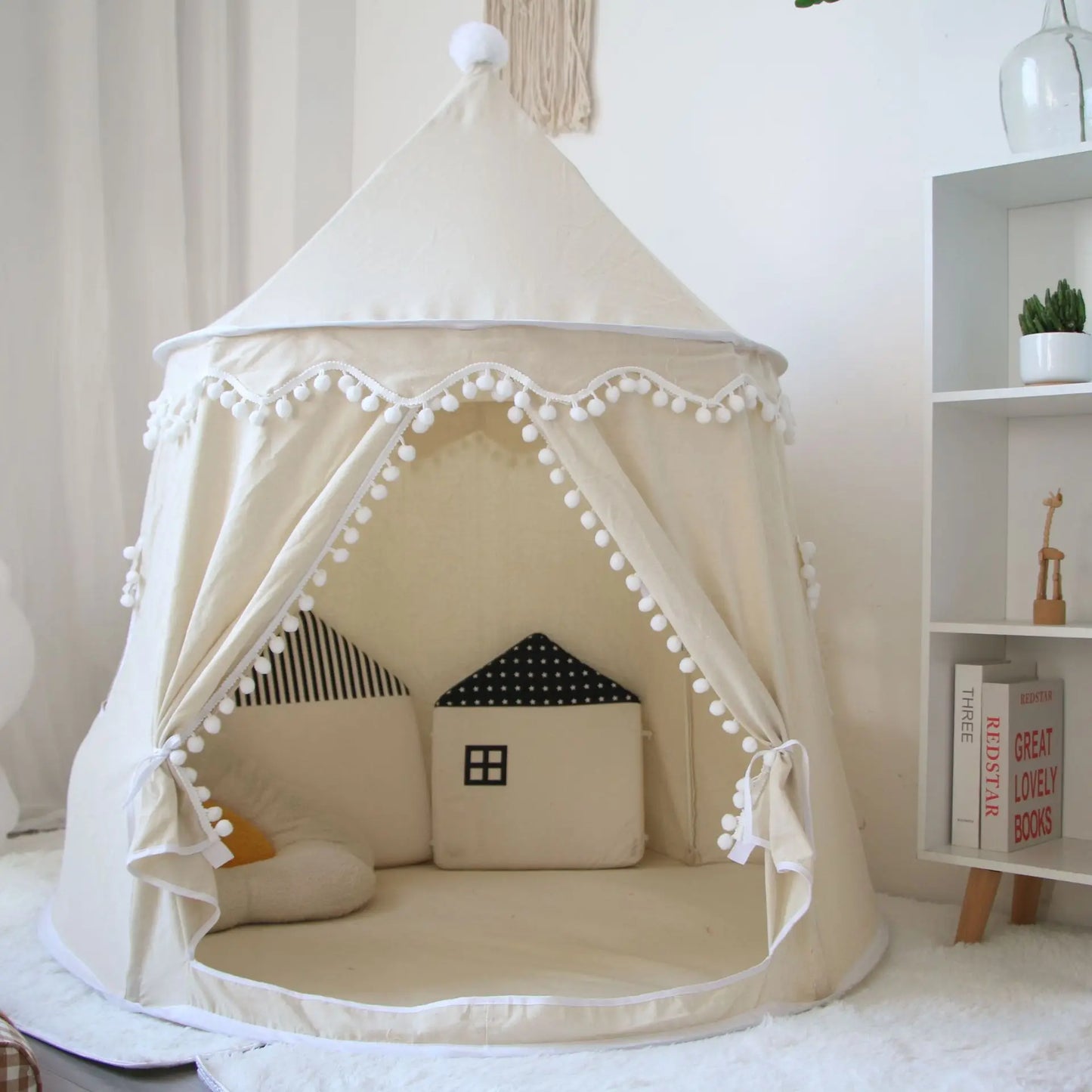 A beige Children's Bali Teepee Tent with a whimsical design, featuring pom-pom details and a small house shape on the front. The tent is set up indoors on a white carpet.