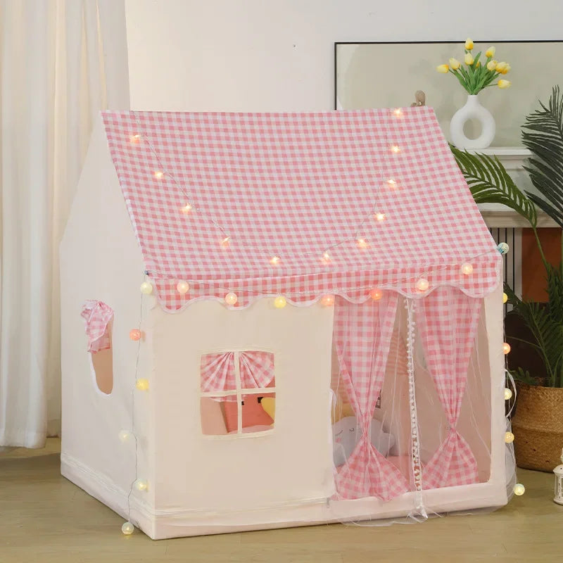 A pink and white gingham playhouse tent with built-in string lights, shown in a styled room setting with a plant and curtain in the background.