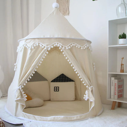 A beige Children's Bali Teepee Tent with a whimsical design, featuring pom-pom details and a small house shape on the front. The tent is set up indoors on a white carpet.