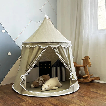 A beige Children's Bali Teepee Tent with a whimsical design, featuring pom-pom details and a small house shape on the front. The tent is set up indoors on a white carpet.