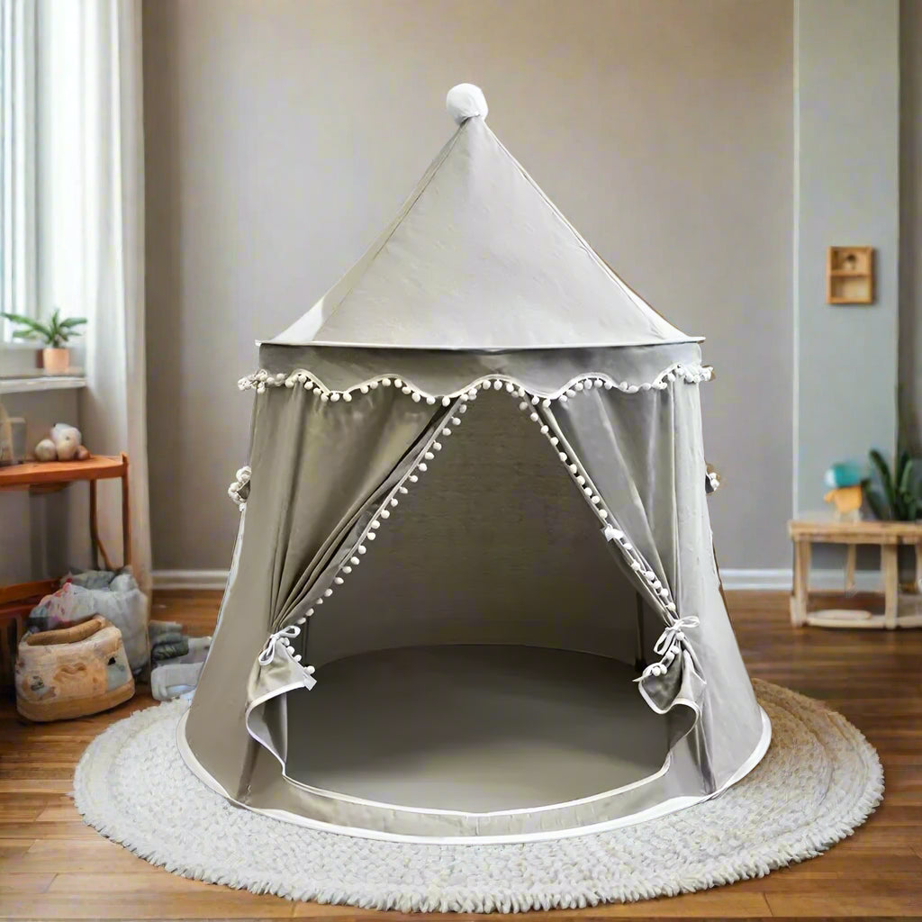A beige Children's Bali Teepee Tent with a whimsical design, featuring pom-pom details and a small house shape on the front. The tent is set up indoors on a white carpet.