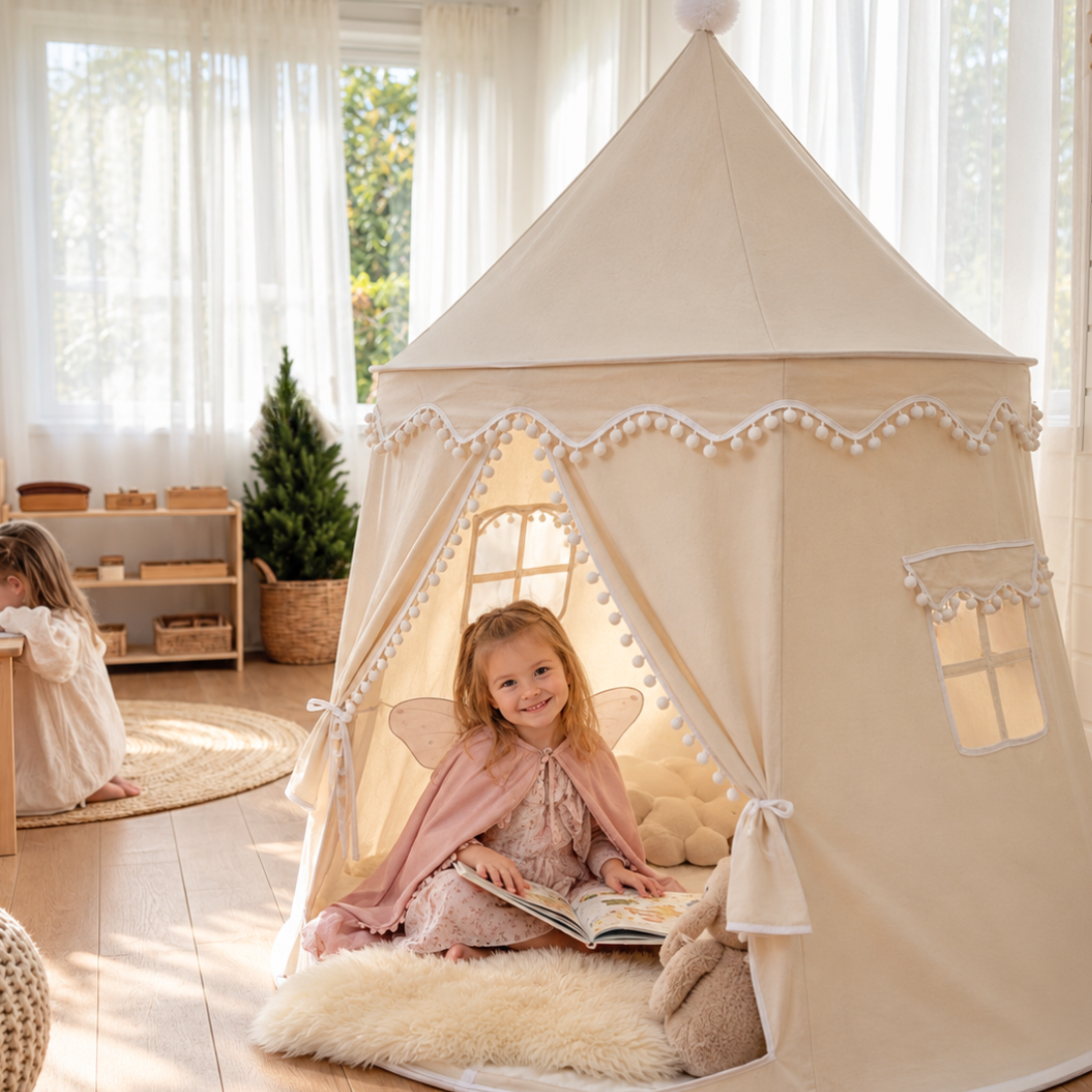 Smiling girl in fairy wings reading a storybook inside the Bali Teepee, a cream pom pom kids play tent, in a sunny playroom